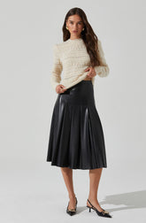 A woman is wearing a cream-colored, long-sleeved crocheted sweater with a textured pattern, paired with a high-waisted, black pleated midi skirt made of a shiny material. She has on black pointed-toe heels with ankle straps and gold details.
